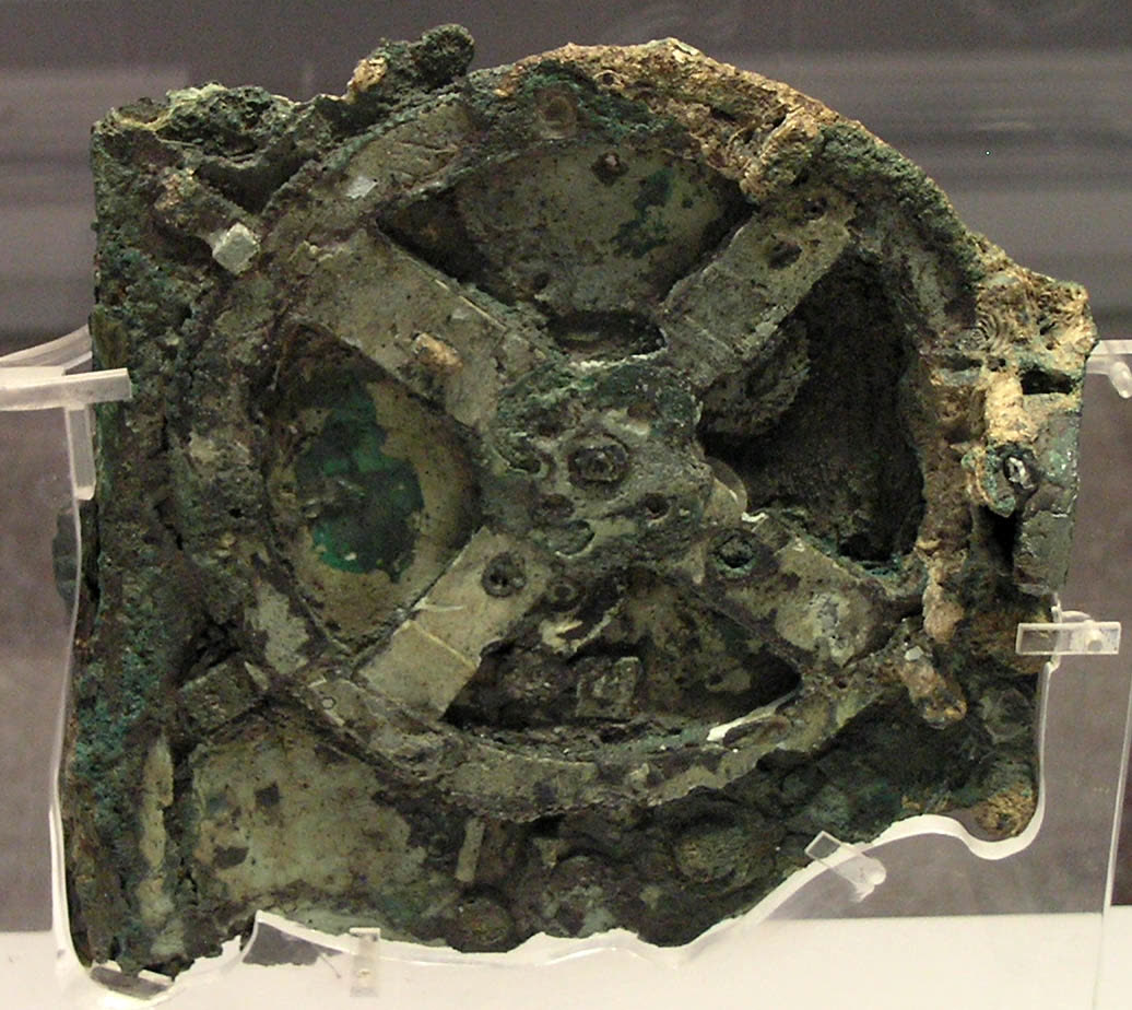 A flat, green-black lump of corroded bronze on a clear stand; a large wheel with four spokes shows in its face