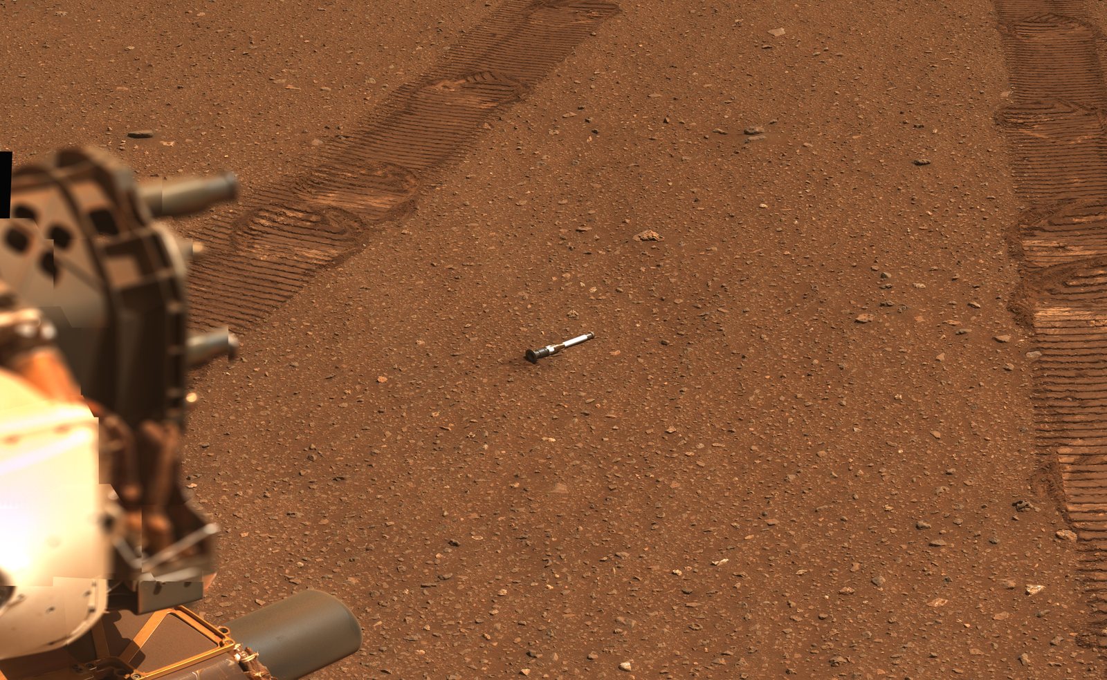 Rover tracks across flat red ground with a small silver tube lying on the sand