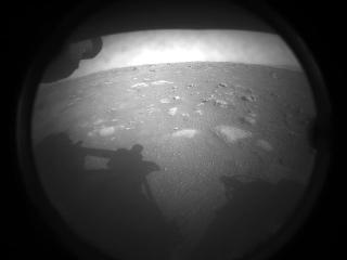 A grainy black-and-white image of the Martian ground seen through a round lens, with the rover’s shadow at the left