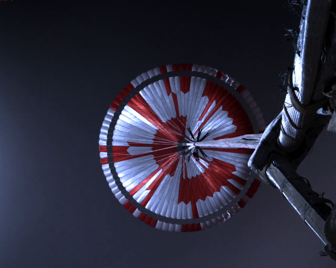 The fully inflated parachute seen from below, red and white panels in a pattern, against a dark sky